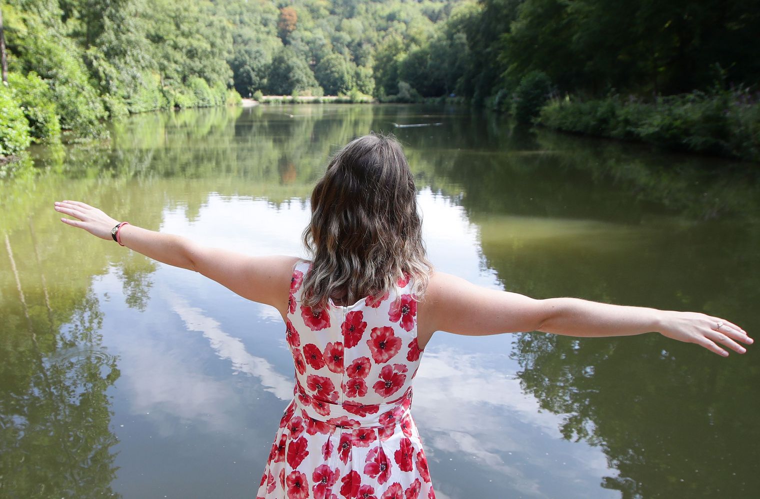 Frau in einem Blumenkleis steht mit horizontal gestreckten Armen vor einem kleinen Waldsee